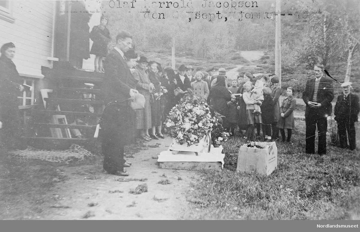 En del folk samlet rundt en kiste.Kisten er pyntet med blomster Bildet er tatt i forbindelse med begravelen til Olaf Harrold Jakobsen, Hamlot, 26. september 1940. Olaf Harrold Jakobsen døde av blindtarmbetennelse 18. september 1940. *