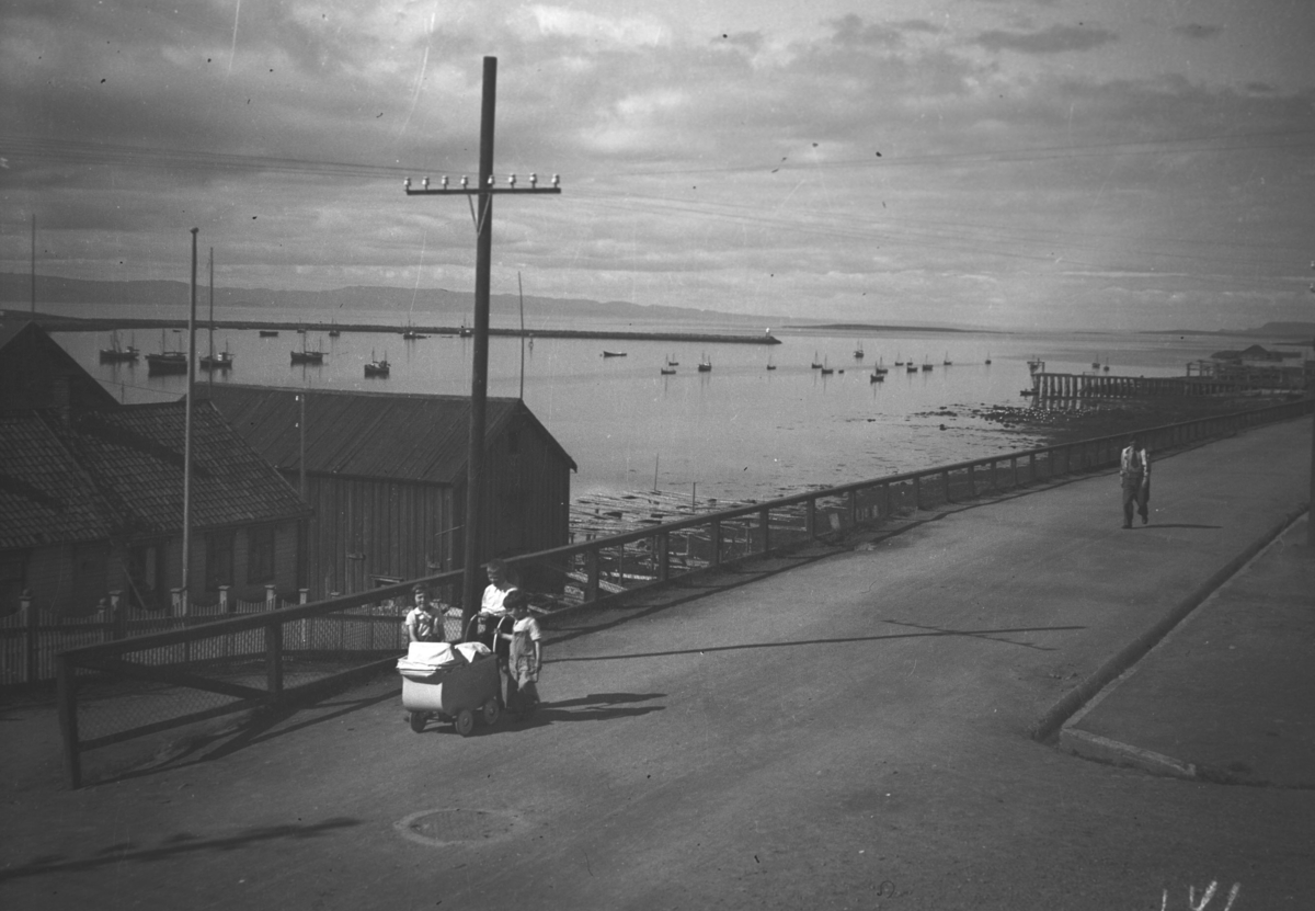 En barnevogn trilles oppover Havnegata, barna bak den er ukjente. Nordahls fiskefabrikk ses til venstre på bildet og Bekola-kaia til høyre. Mange småbåter ligger fortøyd ute i havna.