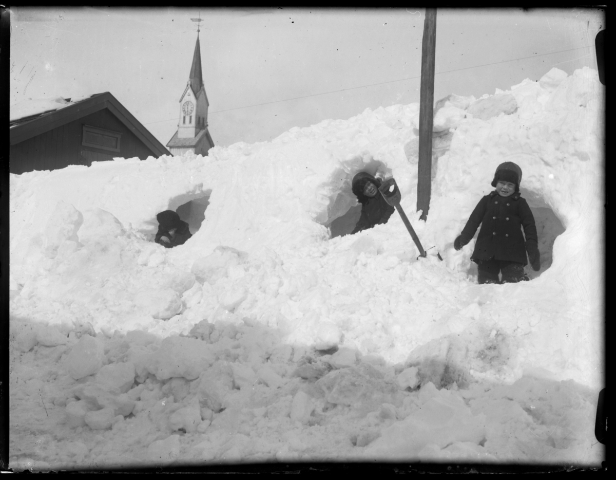 Tre gutter har gravd seg snøhuler i ei fonn, og kikker ut av dem til ære for fotografen. I bakgrunnen bak snøfonnene ser vi taket på husene i prestegården og spiret på kirken