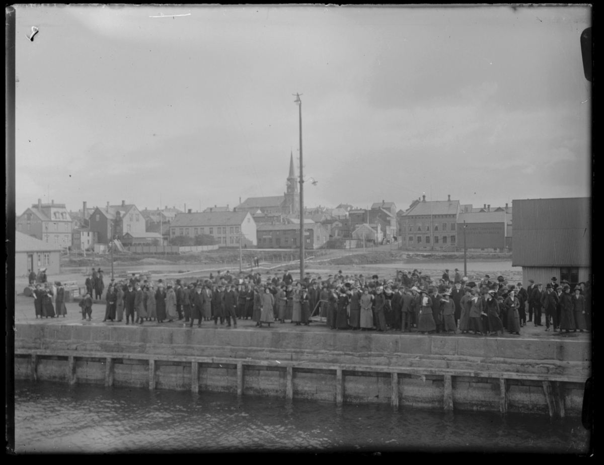 Kaia i Vardø fotografert fra båt, antakelig Hurtigruta. Det har samlet seg en folkemengde på dampskipskaia. I bakgrunnen ser vi byen med bl.a. kirka sentralt i bildet