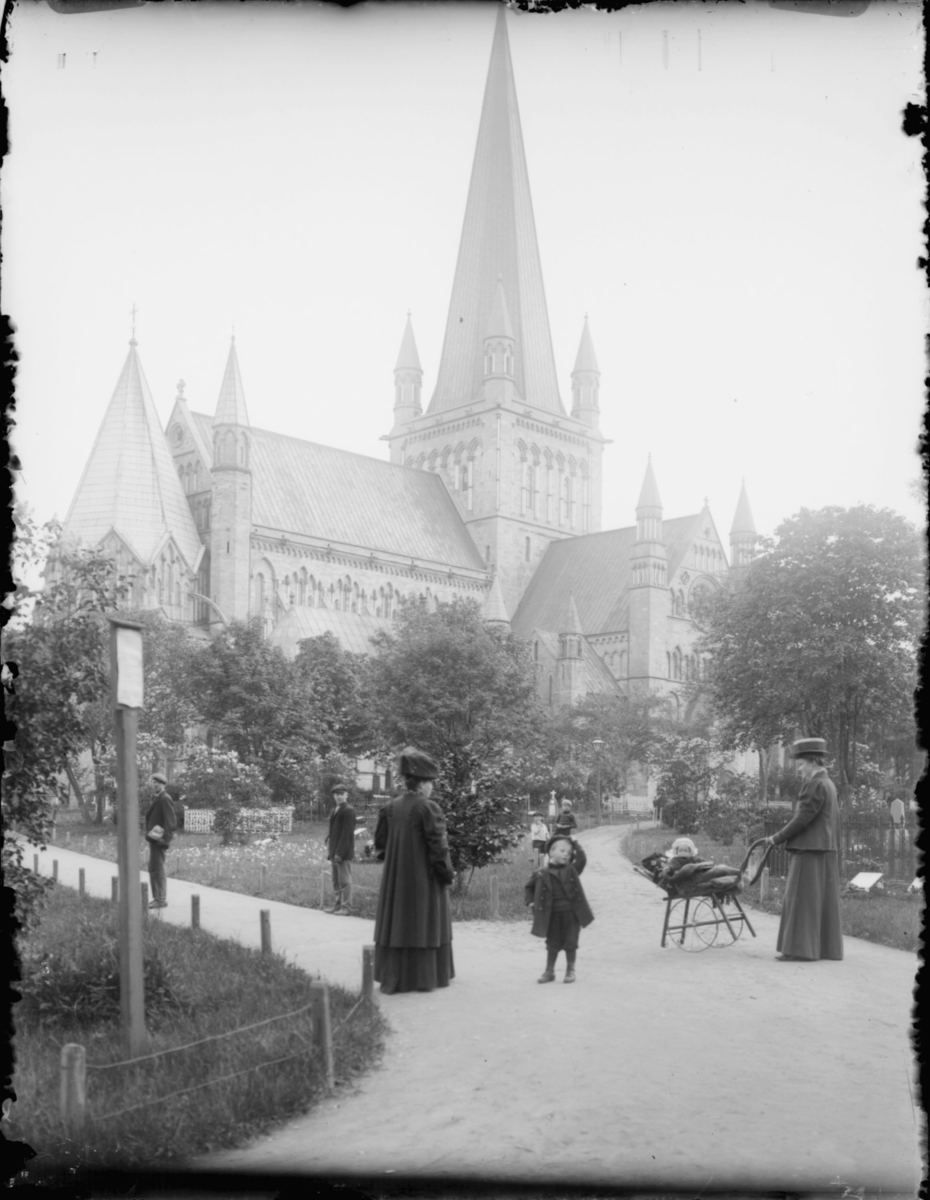 På en gravlund utenfor en kirke. To menn står til venstre. Foran står to kvinner, en med et barn i barnevogn. Tre barn til med på bildet.