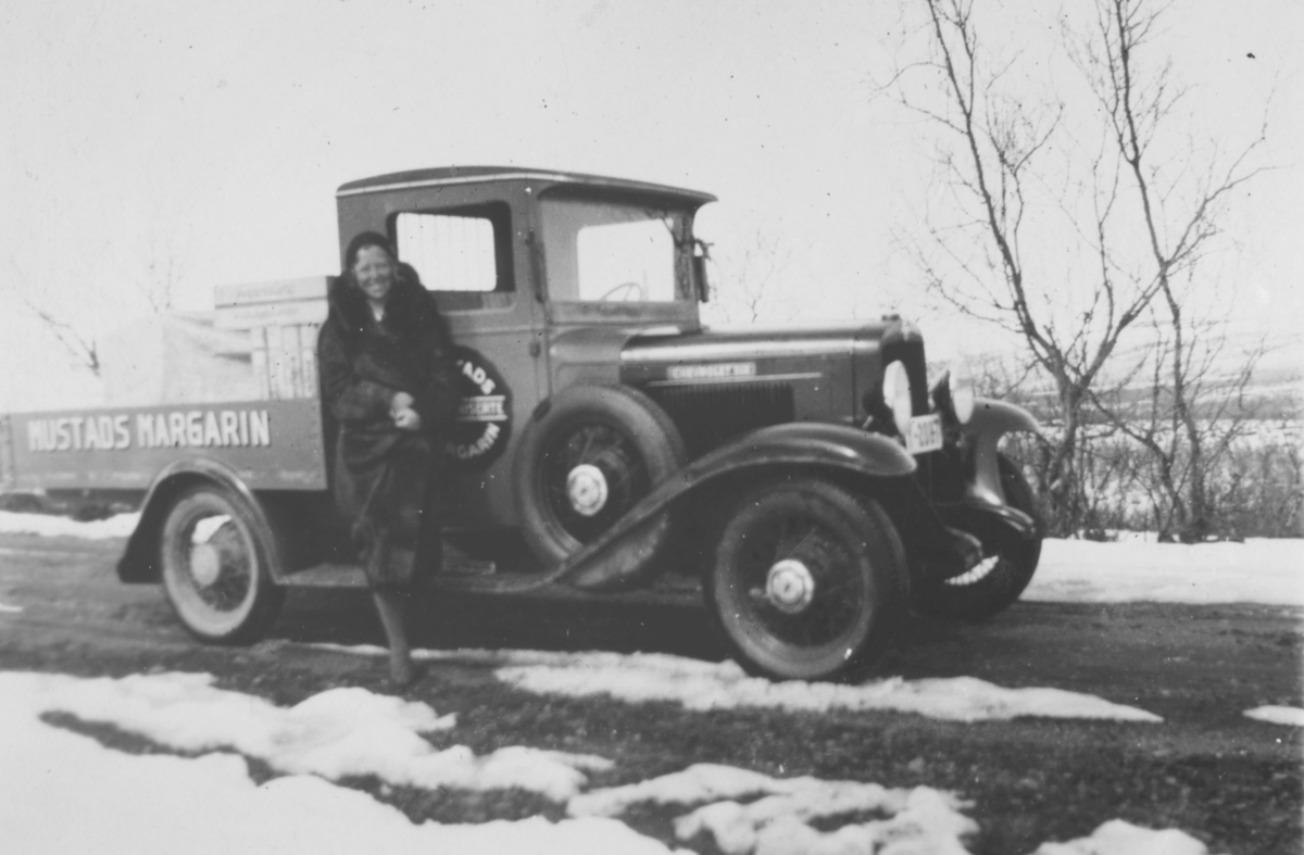 En dame står ved siden av en Chevrolet pickup 1931-33 modell. På lastekarmen står det Mustads margarin. Det er litt snø på bakken. I veikanten et par trær eller busker.