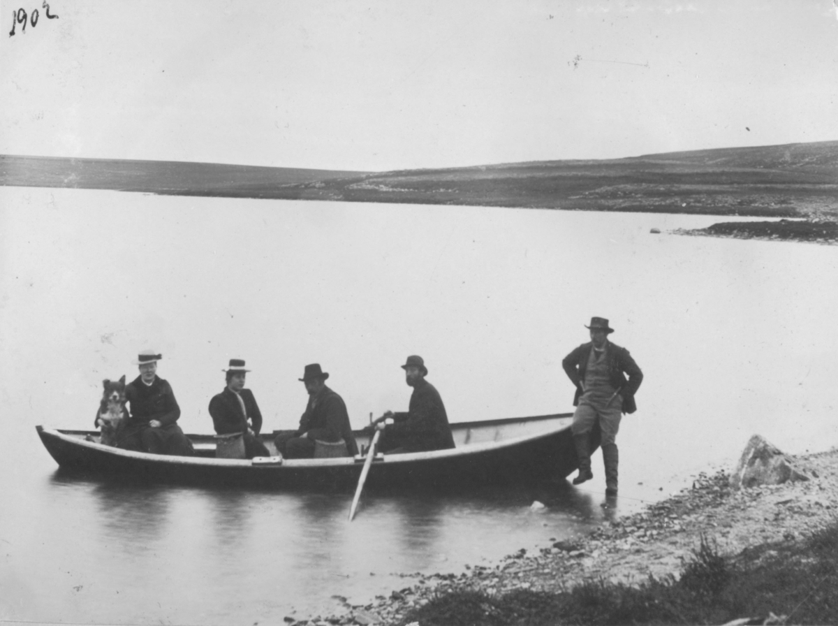 Det sitter 4 stykker i en robåt. To damer og to menn. En tredje mann sitter foran på båten  med en fot i vannet. Damen bakerst i båten har en hund ved siden av seg.