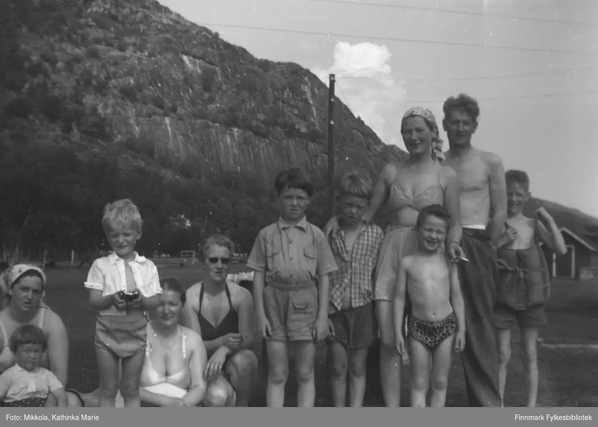 Mikkola-søsknene med barn en fin sommerdag på Mikkelsnes. Alle er lettkledte og flere har badeklær på. Fra venstre: Alfhild Mikkola med datteren Lillian. Sture Olsen Lie, Gudrun Olsen Lie, Herlaug Digre, Roy og Torgeir Mikkola, Kari Havreberg med nevøen Tore foran seg, Mikkel Mikkola og Per Olsen helt til høyre. Per har en slags flytevest på seg. Ut fra barnas alder kan bildet være tatt ca. 1956-1960
