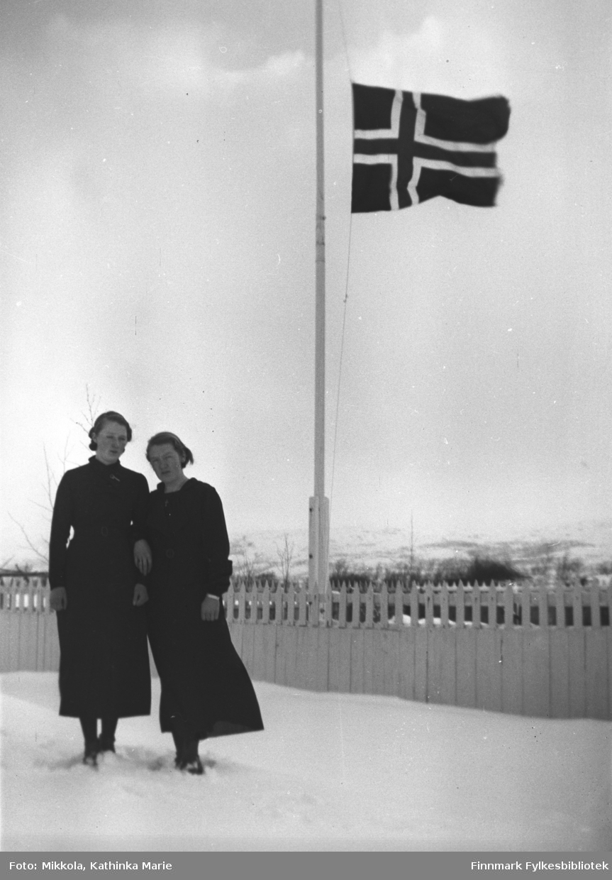 Marine og Synnøve Mikkola i svarte kjoler i snøen på Mikkelsnes. Bak dem vaier flagget på halv stang. Bildet er tatt på begravelsesdagen til jentenes bestemor, Sofie Josefine Mikkola, mor til Aksel Konrad. Hun døde i 1937
