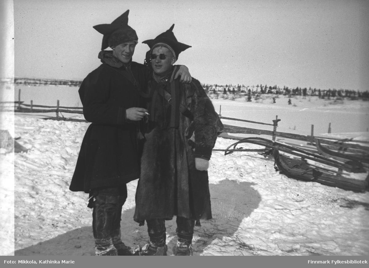 Pakanjoki, ca. 1946-1947? Svogrene Per Sivle Olsen og Storm Mikkola i samekofte og pesk i solskinnet. Per nyter en sigarett. I bakgrunnen to pulker i snøen