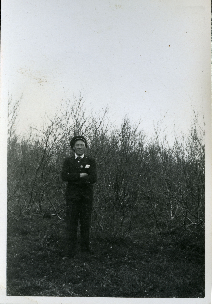 Et fotografi av en mann som muligens kan være Alf Larsen. Alf var arbeider på skiferbruddet i Friarfjord. Her er han kledd i lue, jakke, bukse og slips. Han står foran noen trær.