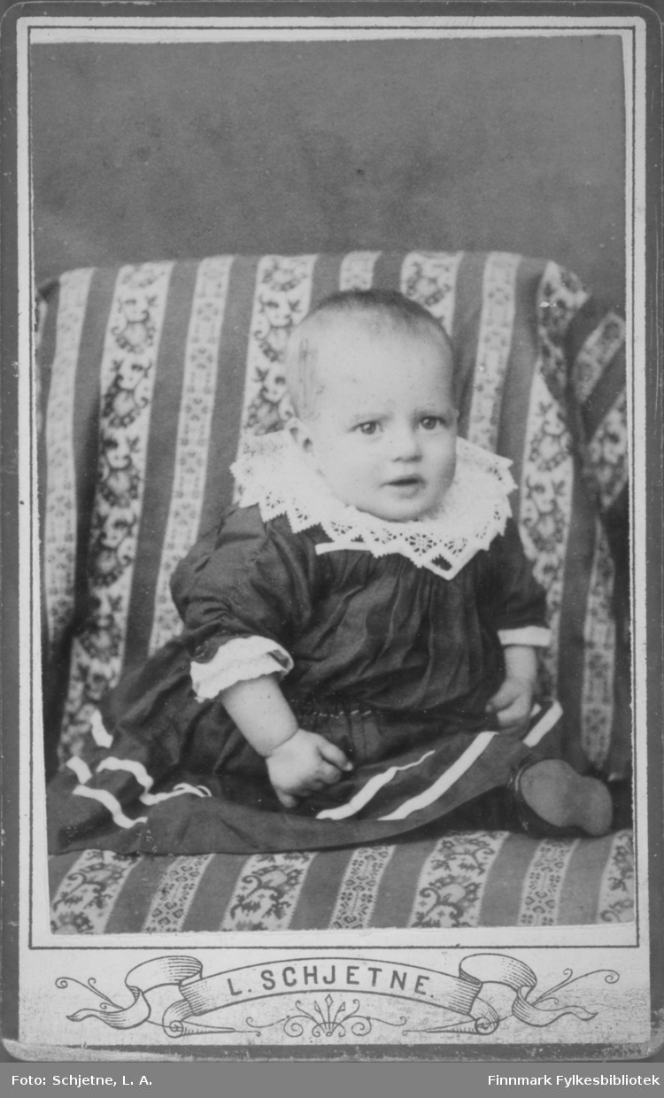 Et lite barn fotografert sittende i en stol med stripet/mønstret trekk. Barnet har en mørk kjole med to hvite striper nede og en bred heklet krage. En sko stikker fram fra kjolen nede til høyre på bildet.