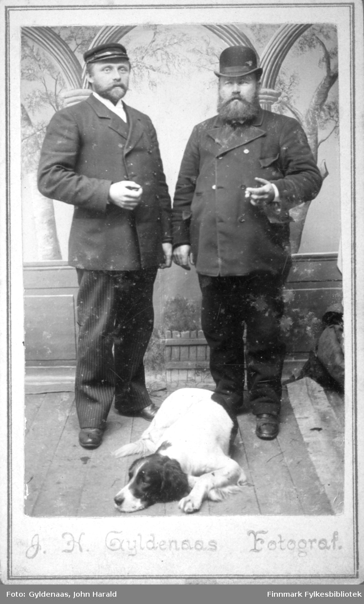 Portrett av to menn. Begge har mørke dresser og hatter på hodet. På mannen til venstre på bildet ses den hvite skjorten i halsen. En sort og hvit hund ligger på plankegulvet foran dem. Et tapet med trær- og bueganger ses på veggen bak dem.