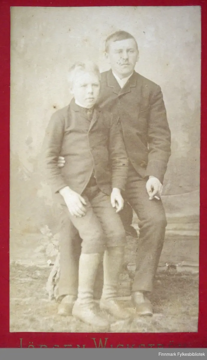 Portrett av to personer. Mannen til høyre på bildet har en mørk dressjakke, mørk bukse og sko. En hvit skjorte eller genser vises i halsen. Gutten til venstre på bildet sitter på kneet til mannen og har en mørk jakke, litt lysere bukse og sko på seg.