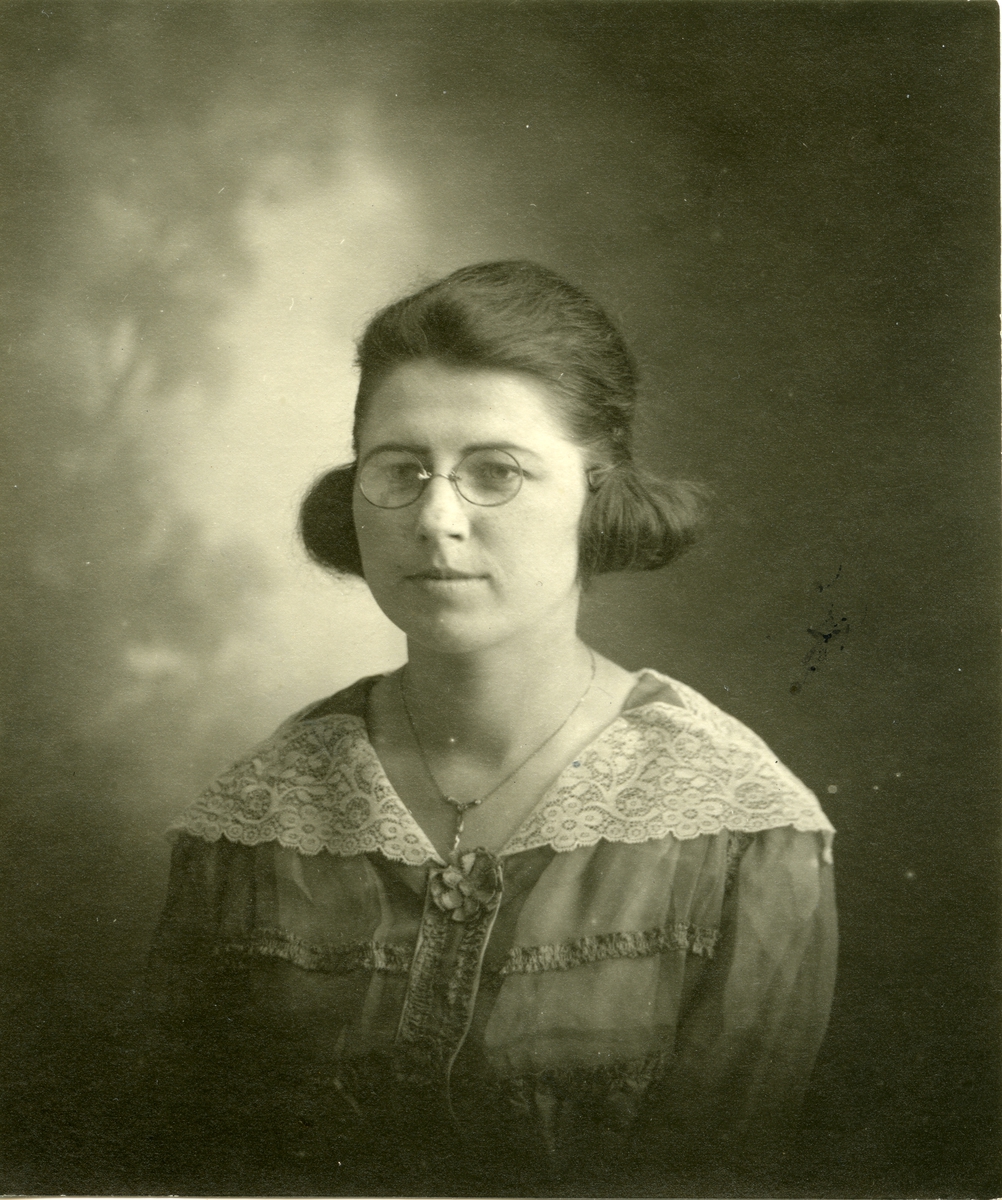 Portrett av kvinne foran lerret. Kvinnen er iført kjole med stor krage og briller.