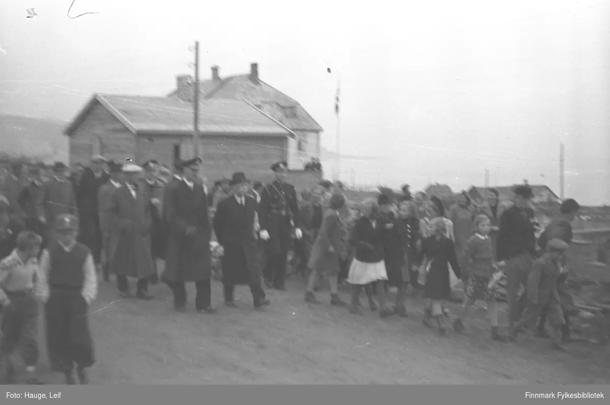 Mye folk står langs veien og ser på mens Kong Haakon og fylkesmann Gabrielsen med følge spaserer. Bygningen i bakgrunnen er kysthospitalet.