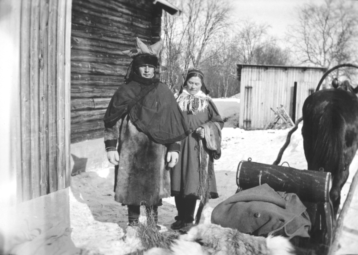 En mann og en kvinne kledt i samiske klær, står utenfor en trebygning. Ved siden av dem står en hest, spent foran en slede. I bakgrunnen er det et vedskjul. Bildet er antakelig tatt innover i Varangerfjorden, eller i Tana