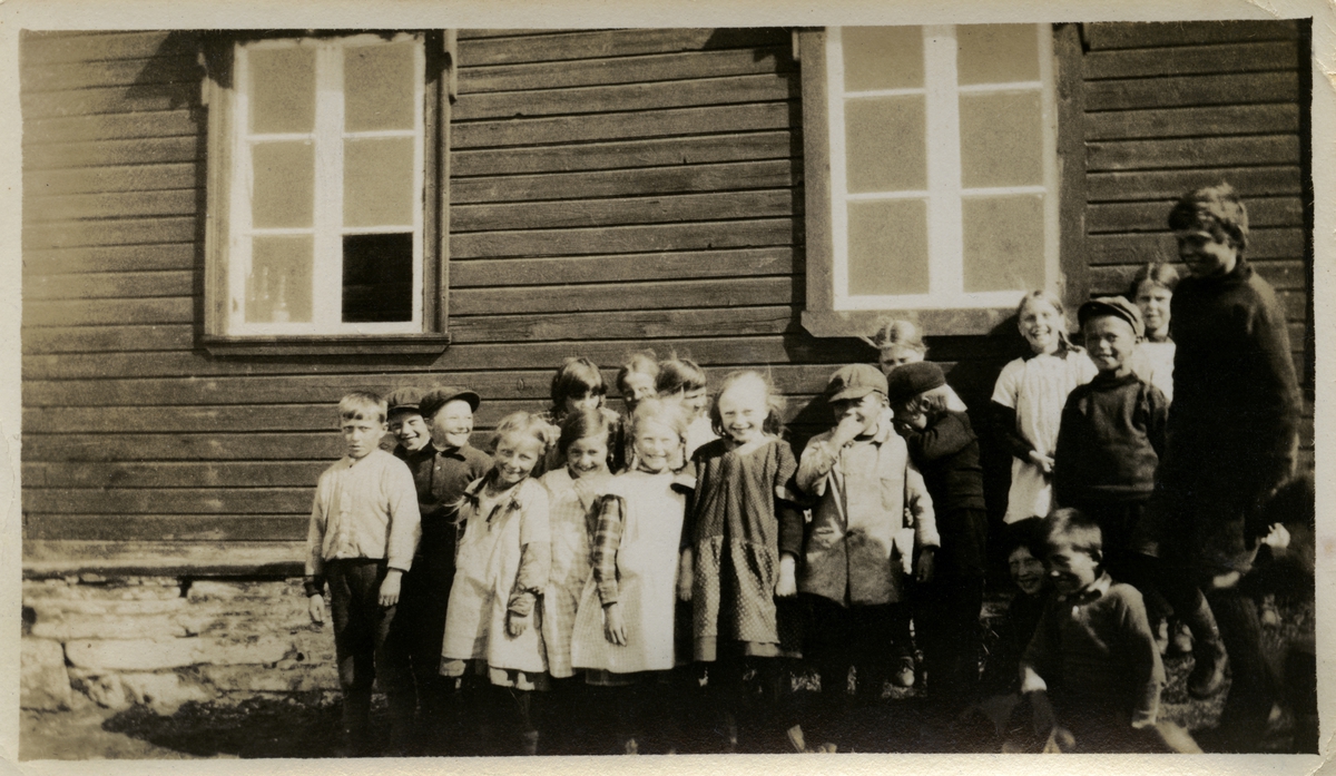 Skoleklasse i Kvalsund skoleåret 1923-24. Smilende jenter er kledd i kjoler, med flettet hår og sløyfer. Guttene har på seg bukser, skjorter, jakker, gensere og luer. Gruppen med skolebarn står foran et hus med grunnmur og vindu.