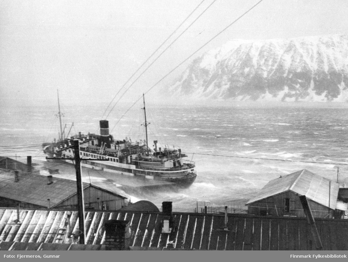 Hurtigruteskipet "DS Lofoten" til kai i Honningsvåg under en storm i mars 1948. Bildet viser tydelig at skipet beveger seg i bølgene selv om det ligger til kai, sjøen har hvite skumtopper og vinden blåser snødrevet rundt i luften.