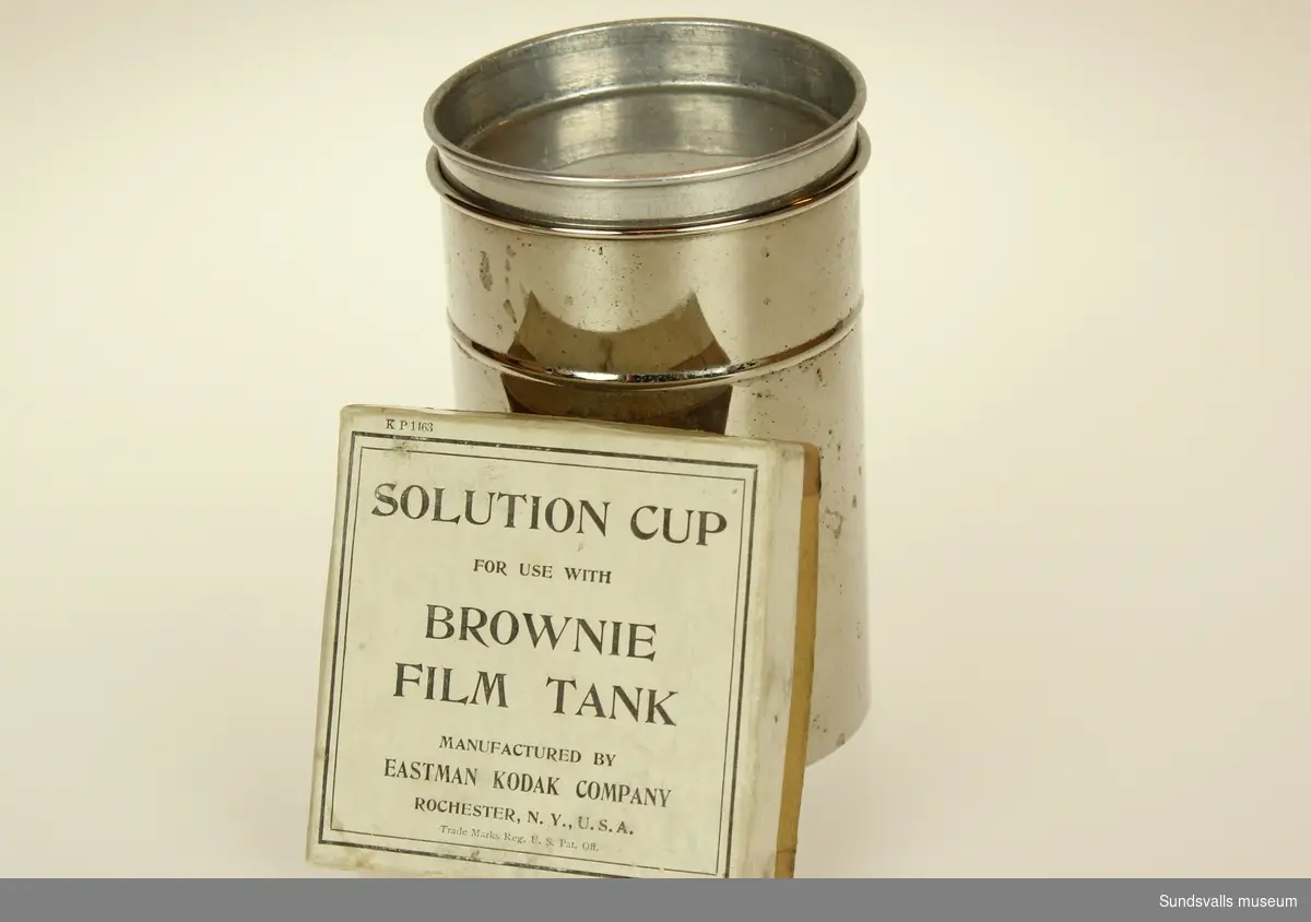 Rund aluminiumbehållare för film. Tillhörande kartong bär texten Solution cup for use with brownie film tank manufactured by Eastman kodak company Rochester N.Y. U. S. A.