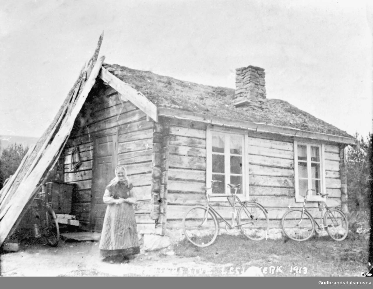 Kvinne foran liten bygning, Magnhilds stue, Lesjaverk, to sykler står blant annet lent inntil veggen. 

