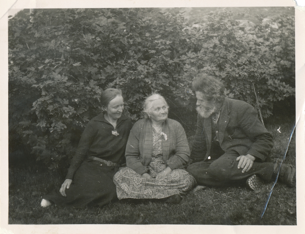 En mann sitter på gresset og snakker med to kvinner. En av de en eldre dame.