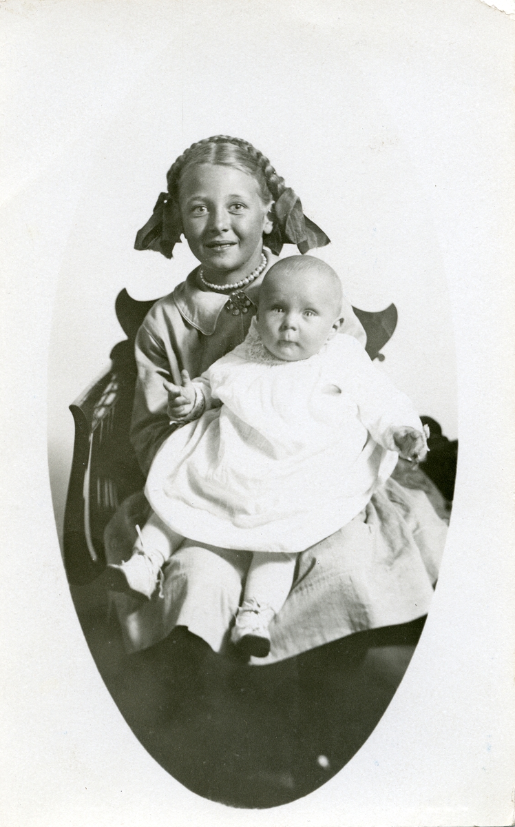 Portrett av to jentebarn foran lerret.En baby og en yngre pike. Jentene er iført finkjoler og den eldste jenten har flettet og oppsatt hår.