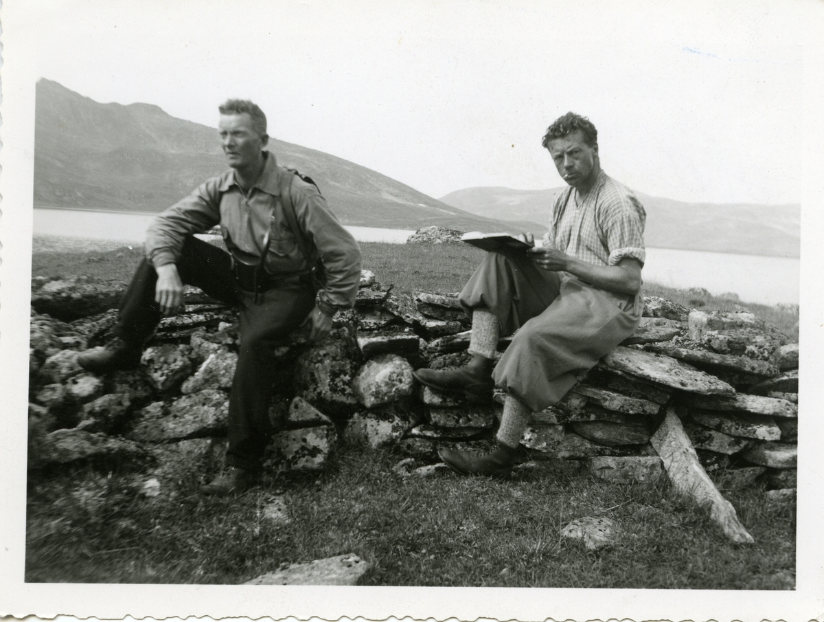 To menn sitter på et steingjerde ved en innsjø. Mennene er iført turklær.