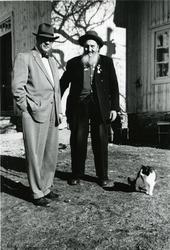 To menn iført hatt står foran noen trehus. Den ene mannen er