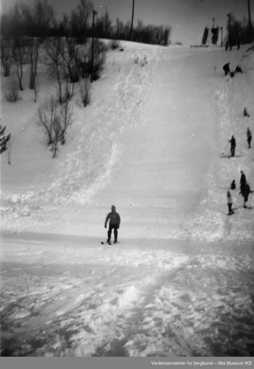 En akebakke med en person som renner ned på ski. Flere mennesker som er på tur opp bakken.