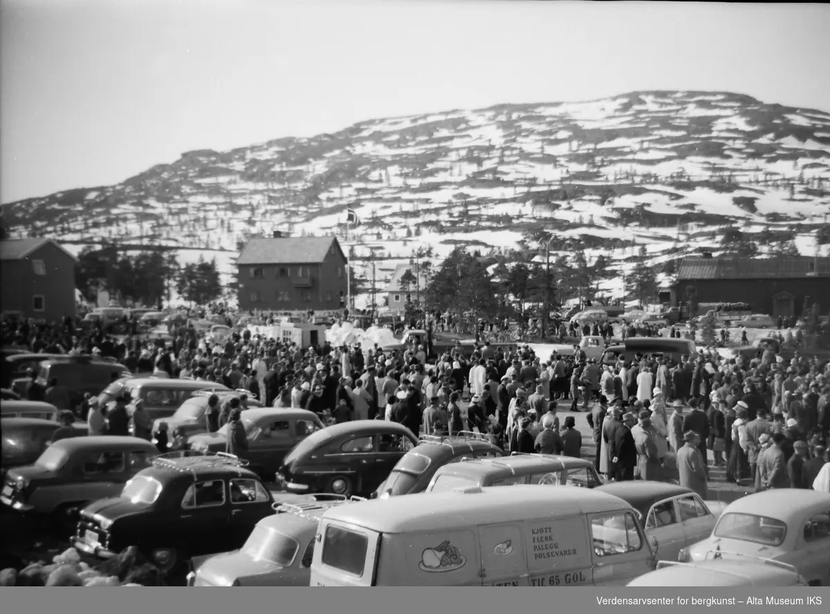 En stor folkemengde har samlet seg på toppen av Bukta-bakken i Alta for å feire 17. mai. Biler står på rekke og rad. Komsatoppen kan ses i bakgrunnen.
Bildet er tatt på våren 1962.