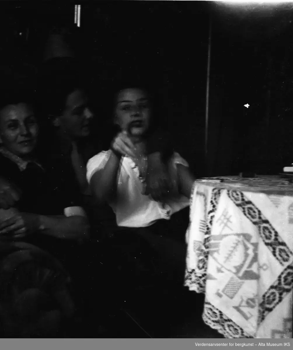 En ung mann og to unge kvinner sitter i en sofa. Mannen har hånden rundt en av kvinnene, som peker mot kameraet.