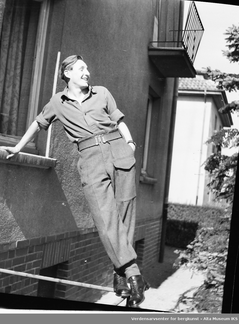 En ung mann i uniform lener seg mot en vinduskarm. Han står over bakken på en tynn jernstang eller et gjerde.