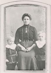 Portrett av en kvinne med finklær og søljer og to barn ståen