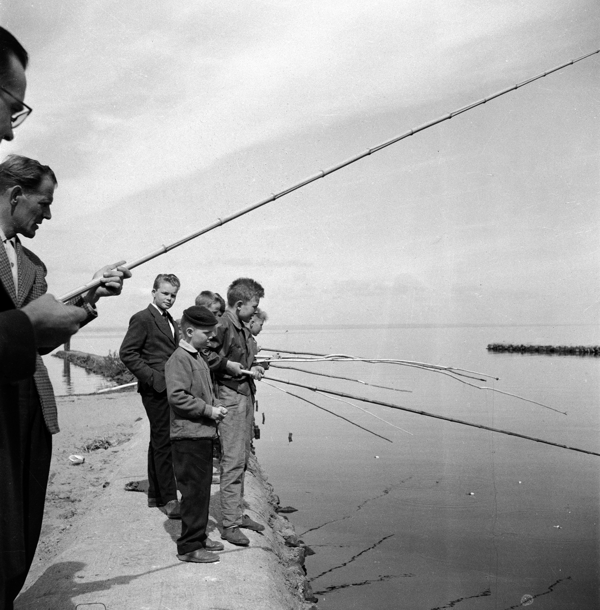 Några ungdomar i Huskvarna står vid Vätterstranden och väntar på att fisken ska nappa. Mannen till vänster med fiskespö är Gösta "Tula" Andersson och till höger om honom står Per Arne Carlman.