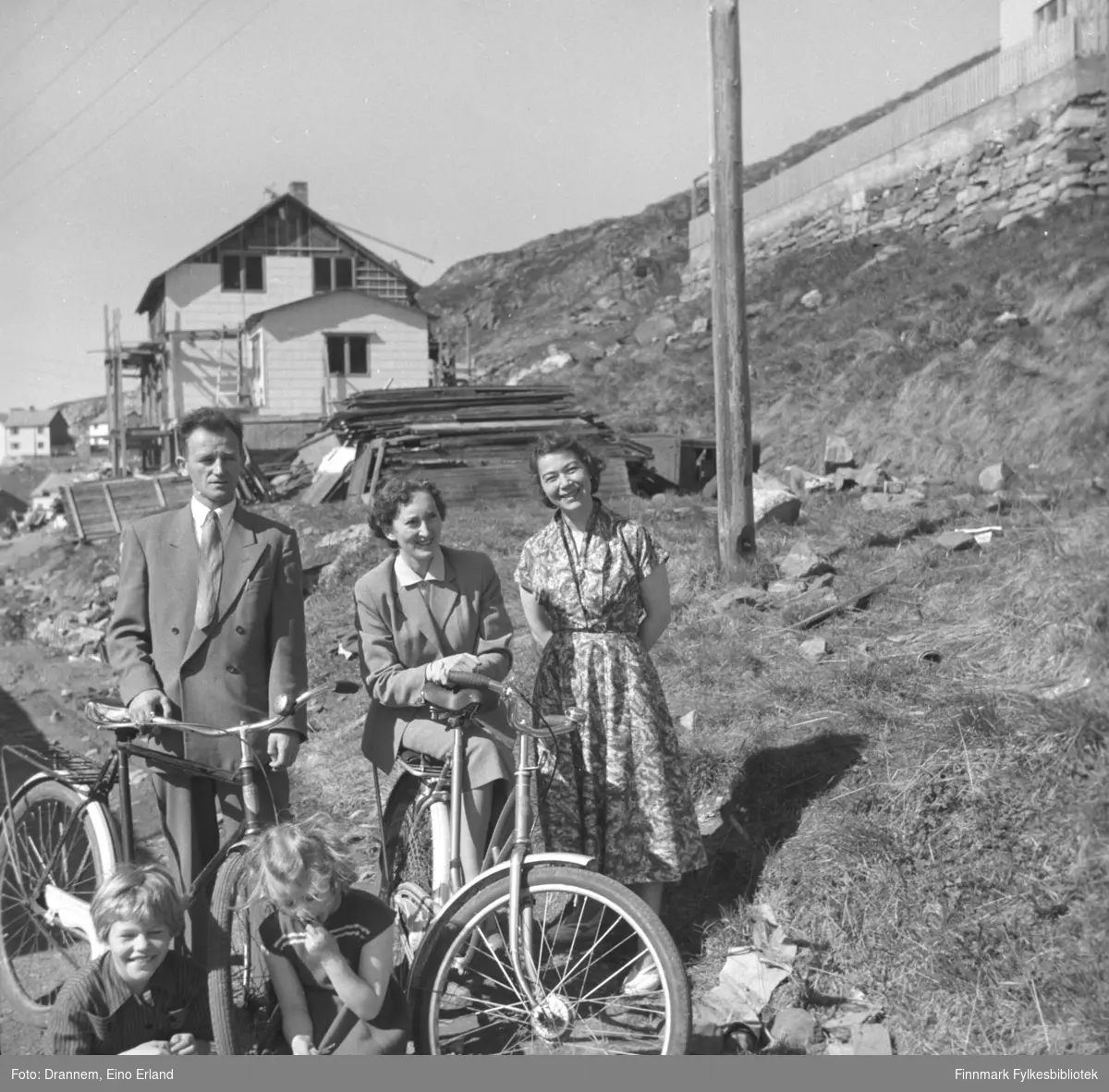 Gruppebilde av smilende mennesker fotografert på veien i Hammerfest. I bakgrunnen ser man et gjenreisningshus under bygging. Barna på bildet heter Vigdis Hansen og Turid Drannem, paret med sykler er ukjente og helt til høyre er Turid Drannem