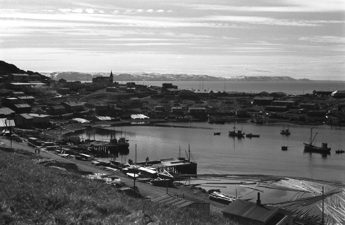Bebyggelsen reises i Honningsvåg etter andre verdenskrig. I forgrunnen ligger byggematerialer og flere båter ligger for anker  i havna.

Arkitekt Ola Hanche-Olsen arbeidet ved Brente Steders Reguleringskontor i 1946. Hovedadministrasjon for gjenreisning av Nord-Troms og Finnmark ble lagt til Harstad og fikk navnet Finnmark kontoret. Landsdelen Nord-Troms og Finnmark blev oppdelt i syv distrikt med hver sin administrasjon. Honningsvåg, distrikt IV, skulle betjene Nordkapp, Lebesby, Porsanger og Karasjok kommune.

Ola Hanche-Olsen har tatt bildene. Han var født 13. mars 1920 i Borre, død 11. februar 1998 i Gjettum. Han var både arkitekt og barnebokforfatter. Han hadde artium fra 1939, arkitekteksamen fra NTH 1946 og arbeidet deretter ved Finnmarkskontoret 1946–48 før han etablerte egen arkitektpraksis. Han debuterte som barnebokforfatter i 1974 med lettlest-boka "Knut og sjørøverne", og skrev i alt 12 bøker. Han var XU-agent 1944-45, og var også en aktiv fjellklatrer og friluftsmann. Ola var gift med Solveig Hanche-Olsen (f. Falkenberg); de fikk 3 barn, blant dem matematikeren Harald Hanche-Olsen.

