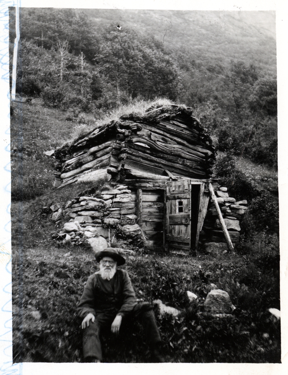 Eldre mann med hvitt skjegg sitter utenfor en gammel, falleferdig låve og fjøs, bygget i bratt terreng.