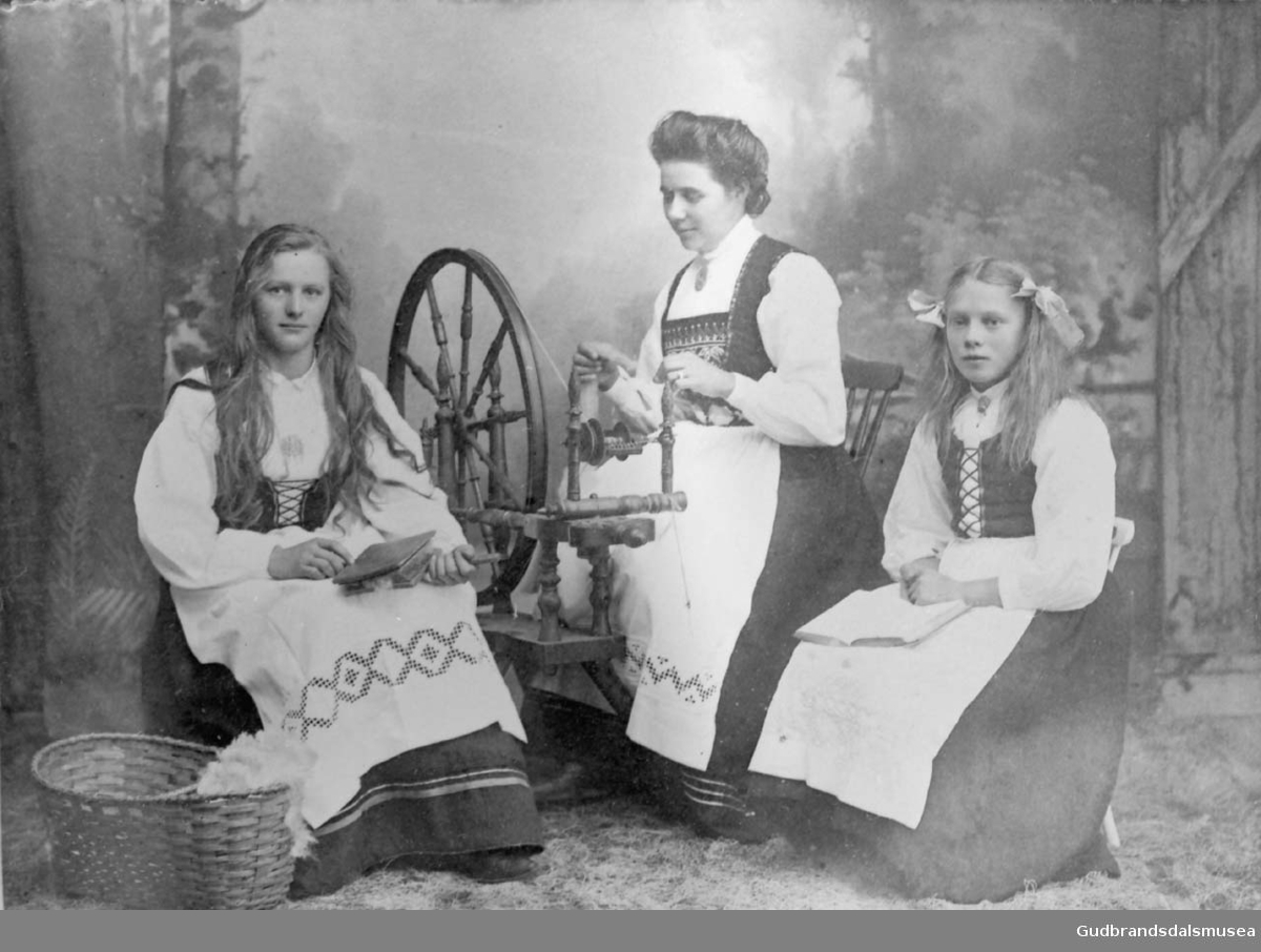 Portrettbilde, tre personer i atelier, kvinne spinner på rokk, en jente karder ull, en jente sitter med bok på fanget. 