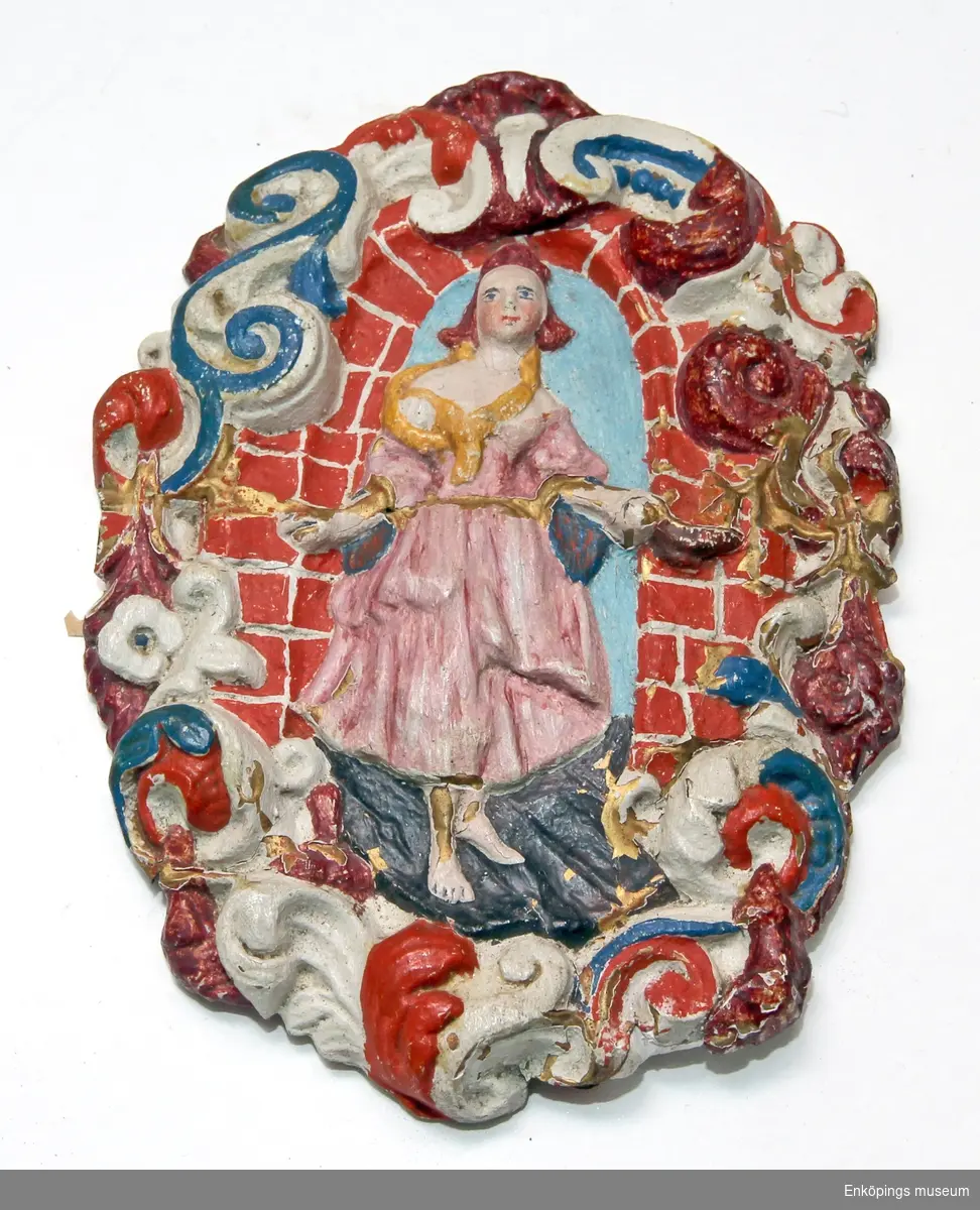 Ornament av gips med målad polykrom dekor reliefmönster föreställande kvinnogestalt i helfigur i ett tegelvalv, ram bestående av rokokoornament. På baksidan rester av målning huvudsakligen i gult, bild av 2 st kyssande änglar samt text.