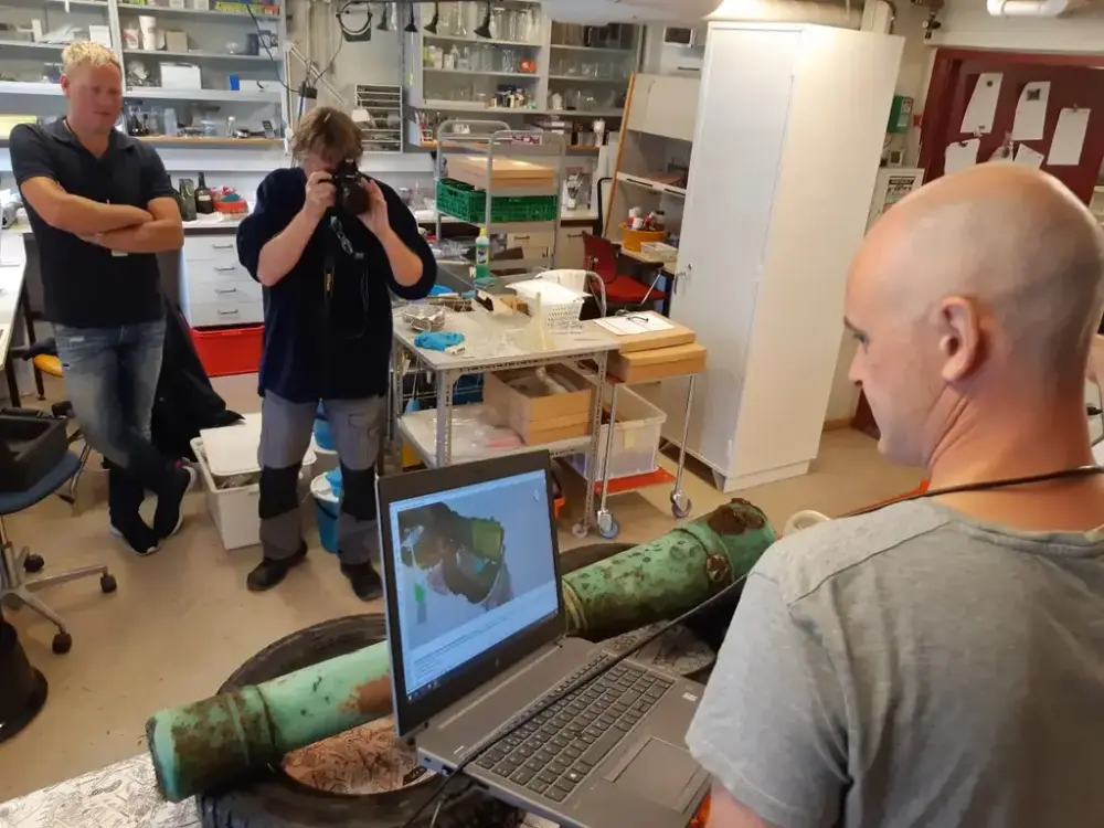 Kanon ligger polstret med bildekk på et bord i et laboratorium. En person holder en bærbar datamskin tilkoblet en håndholdt skanner og måler inn kanonen i 3D. En person fotograferer kanonen fra motsatt side.