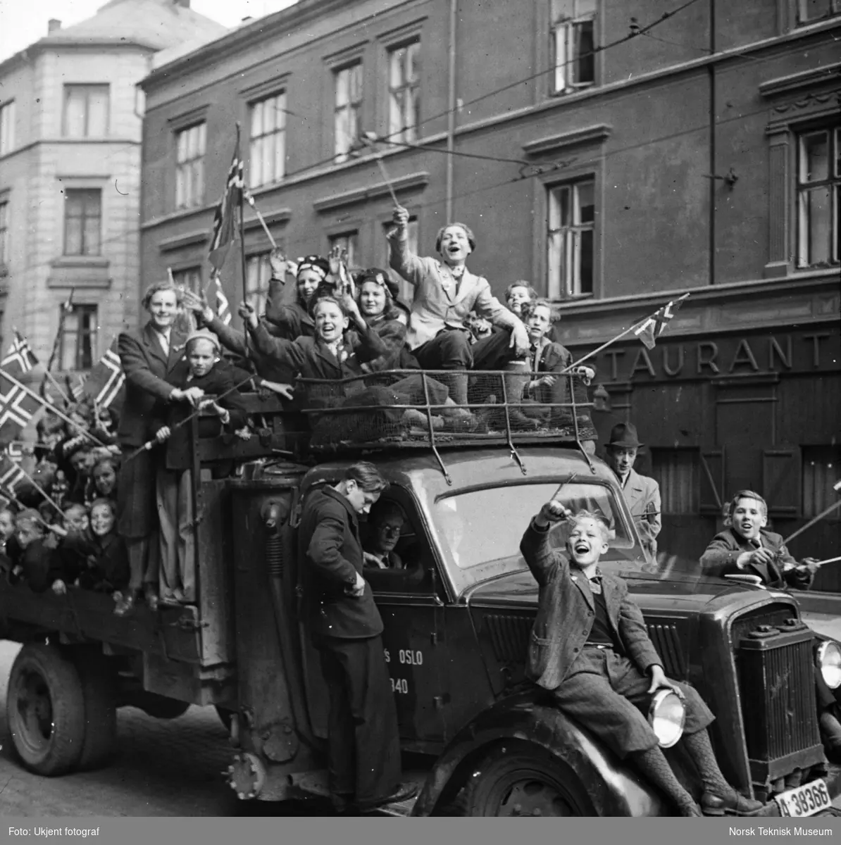 Jublende kvinner og barn med flagg på lasteplanet og panseret til en lastebil med registreringsnummeret A-38366 kjører gjennom Oslos gater, mai 1945,