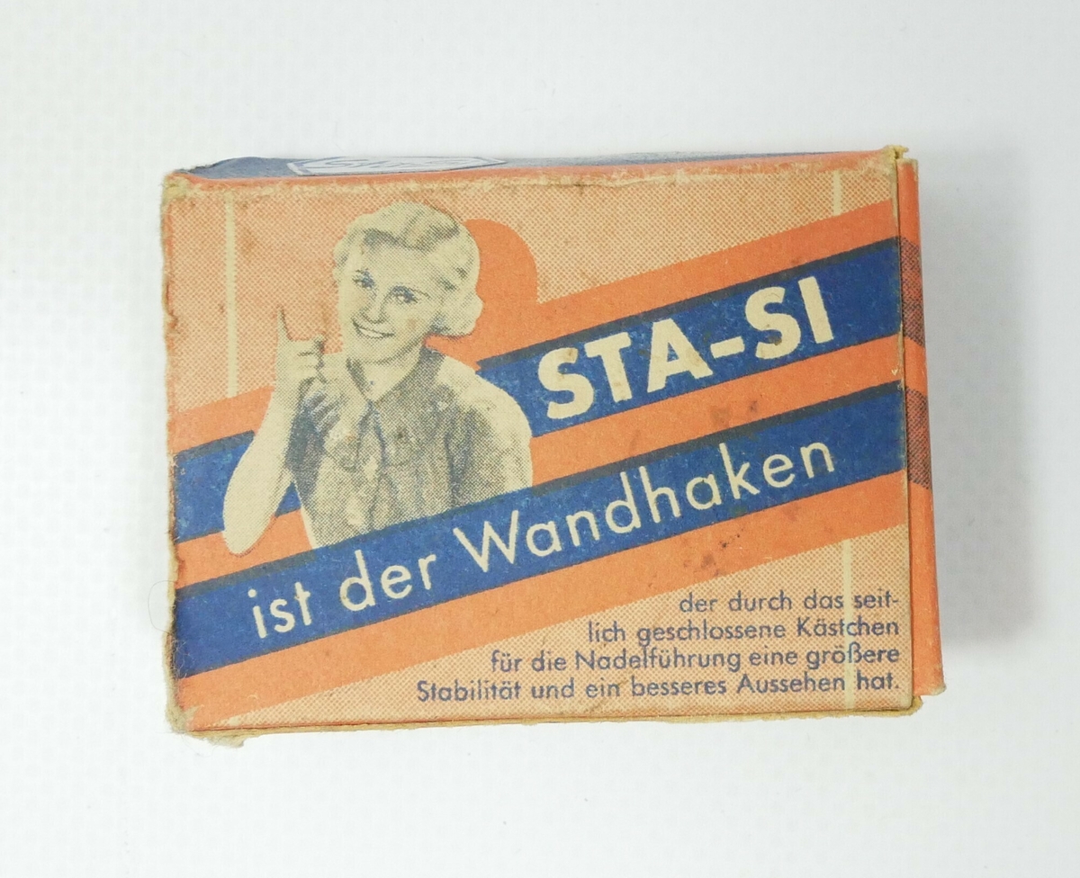 Rektangulær kartongemballasje i rødt og blått med påskrift på tysk. Emballasjen har inneholdt veggkroker. Den ene kortsiden er åpen og emballasjen er tom. 

Påskrift: 
STA-SI WANDHAKEN // Sta - Si Wandhaken werden nur mit // la gerhärteten Stahlnadeln geliefert 

Påskrift: 
[på tysk]