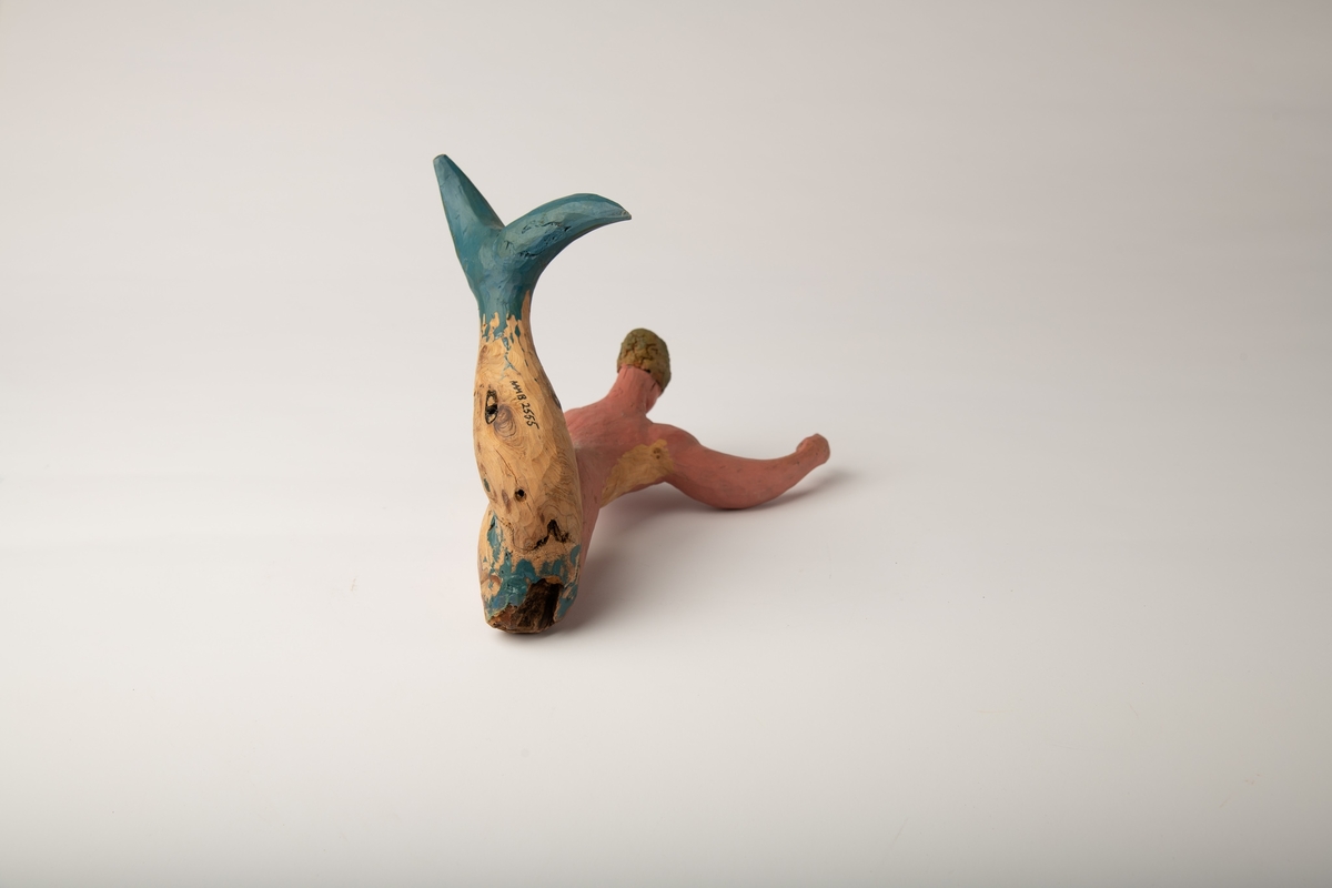 Mannsfigur i vinkel med blå halefinne og delvis rosa kropp. Hendene ut fra kroppen. Skåret av et stykke tre, der treets naturlige form har blitt en del av skulpturens form.