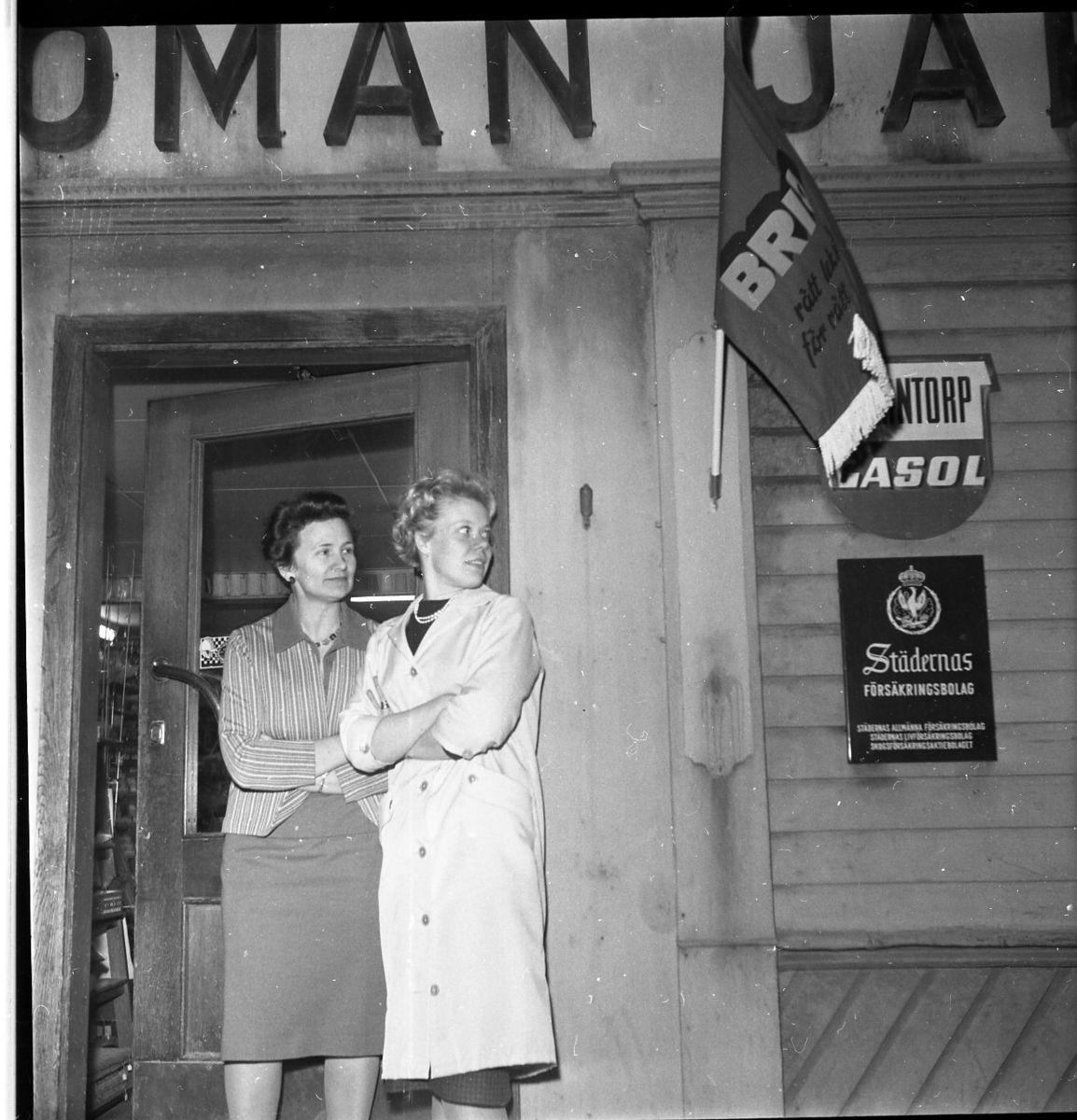 Två kvinnor står i butiksdörren till Romans Järn. Kvinnan till höger (Marianne Rundqvist) har en tunn ljus rock på sig och jobbar troligen i butiken. De tittar åt och höger och väntar sannolikt på Gränna scoutkårs lucia med följe som åker med en VW-pickup.
