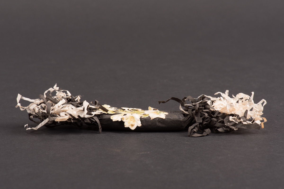 Platt begravningskaramell inlagd i svart silkesppapper med klippta och krusade fransar i svart och vitt i bägge ändarna.
På ovansidan dekorerad med ett påklistrat bokmärke i form av liljor.
