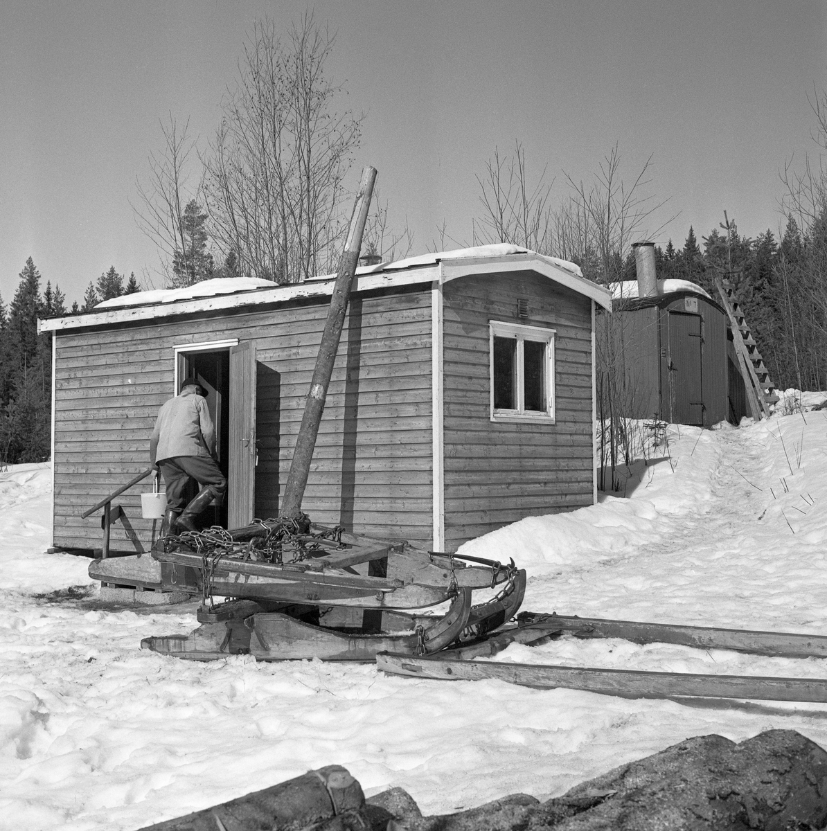 Tømmerslede på snødekt mark framfor flyttbar koiebrakke og stall i Stavåsen i Elverum, Hedmark. Fotografiet blew tatt vinteren 1971, da Jørgen Larsen fra Hernes kjørte tømmer i dette området. Sleden var en såkalt «rustning» (også kalt «reitdoning» eller «bukk og geit»), altså en todelt sledetype. Når det var lass på rustningen hang baksleden («geita») bak forsleden («bukken») i en Y-formet kjetting («trossa»). Den todelte sledeløsningen og den dreibare «banken» (tverrtreet som bar lasset) på forsleden gjorde rustningen til et smidig transportredskap på svingete og staupete veger i skogsterrenget. Når doningen skulle kjøres «tomreipes» tilbake mot hogstteigene hvor det var mer tømmer å hente, var det vanlig å gjøre som Jørgen Larsen hadde gjort her, nemlig å legge geita oppå bukken med en høysekk som sitteunderlag på den øverste sleden. Akkurat da dette fotografiet ble tatt var Jørgen Larsen på veg inn i den brakka han var innkvartert i mens han kjørte tømmer i dette området. Det dreide seg om en rektangulær bindingsverkskonstruksjon med dør på langveggen og vindu i gavlveggen. I bakgrunnen (ovenfor) ser vi også en del av stallbrakka. Begge disse husværene var flyttbare i den forstand at de hadde hjul og kunne trekkes bak en traktor til et annet driftssted på skogsbilveger i barmarkssesongen.