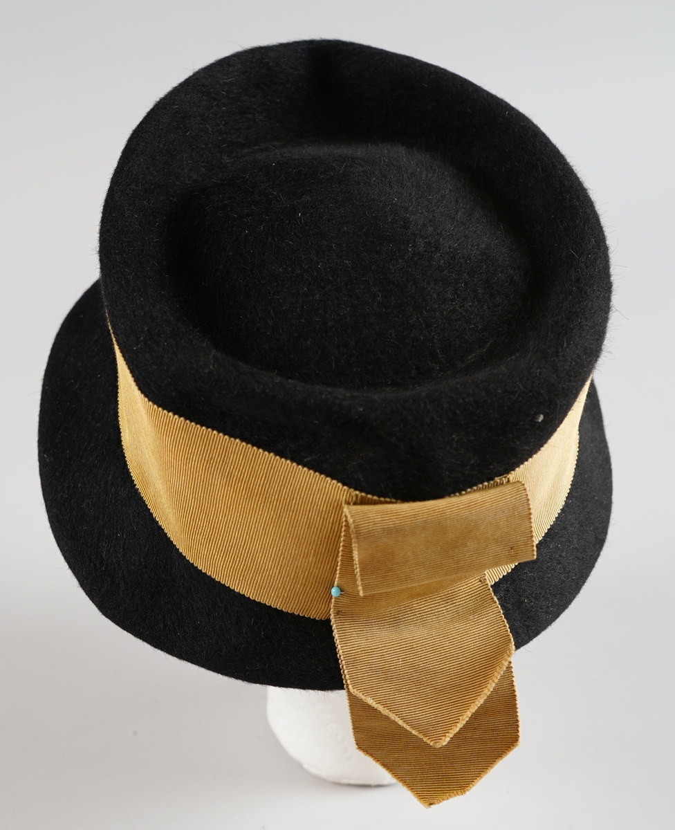 Svart hatt i ullfilt med gyllent pynte band og tynt strikk til feste.