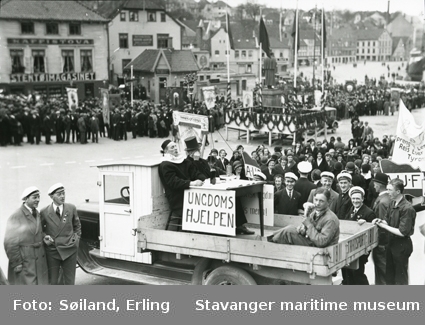 1. mai tog i Stavanger 1932. Folk stilt opp på torget i Stavanger. Foran i motivet er en lastebil med tre utkledde ungdommer på lasteplanet. På en plakat står "Ungdomshjelpen". I bakgrunnen et banner med "AUF".