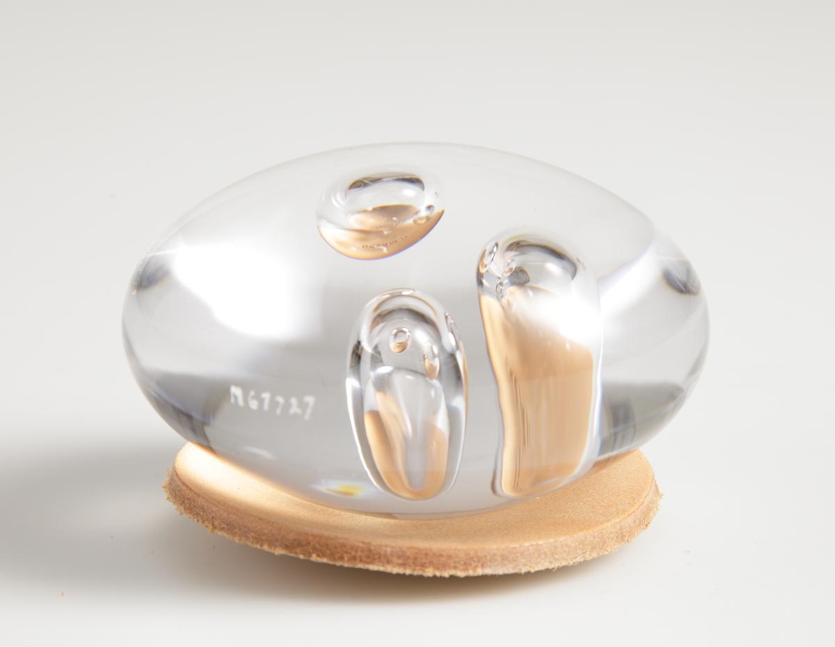 Mindre ovalt glasblock smyckat med tre blåsor inne i glaset.  Blocket ligger på en rund läderbit med texten "DREAM IT".
Etikett: Ofärgad med svart text "M Målerås Sweden Since 1890"