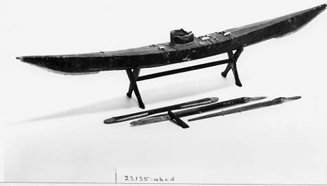 Fartygsmodell av en enmanskajak tillverkad av skinn spänt på trästomme. Till modellen finns även två fångstspjut, paddel och skrå.