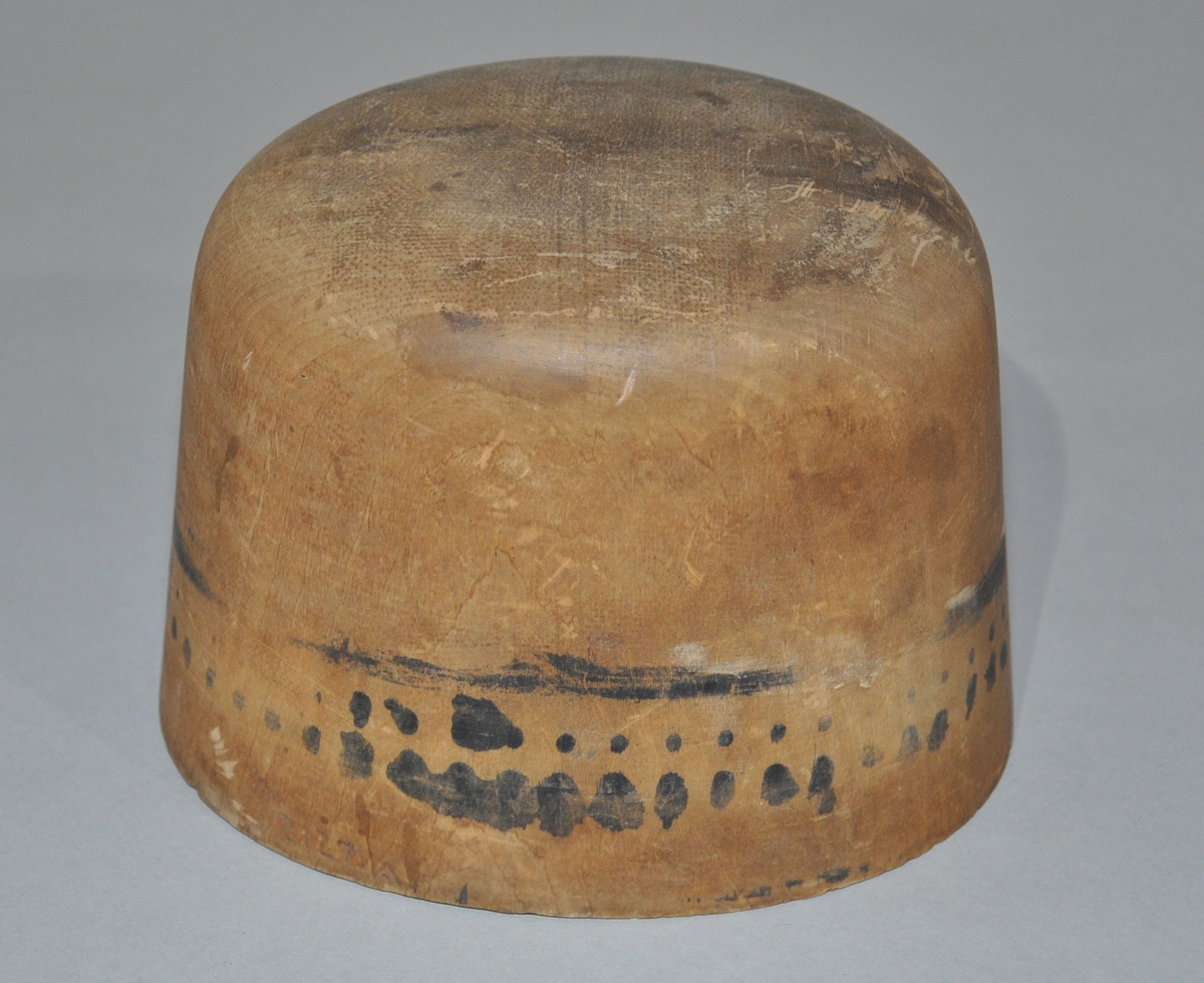 Hatteblokk til pull, av tre. Oval form, med avrundet topp. Hatteblokken har rester av blekk eller annet langs kanten på bunnen.
