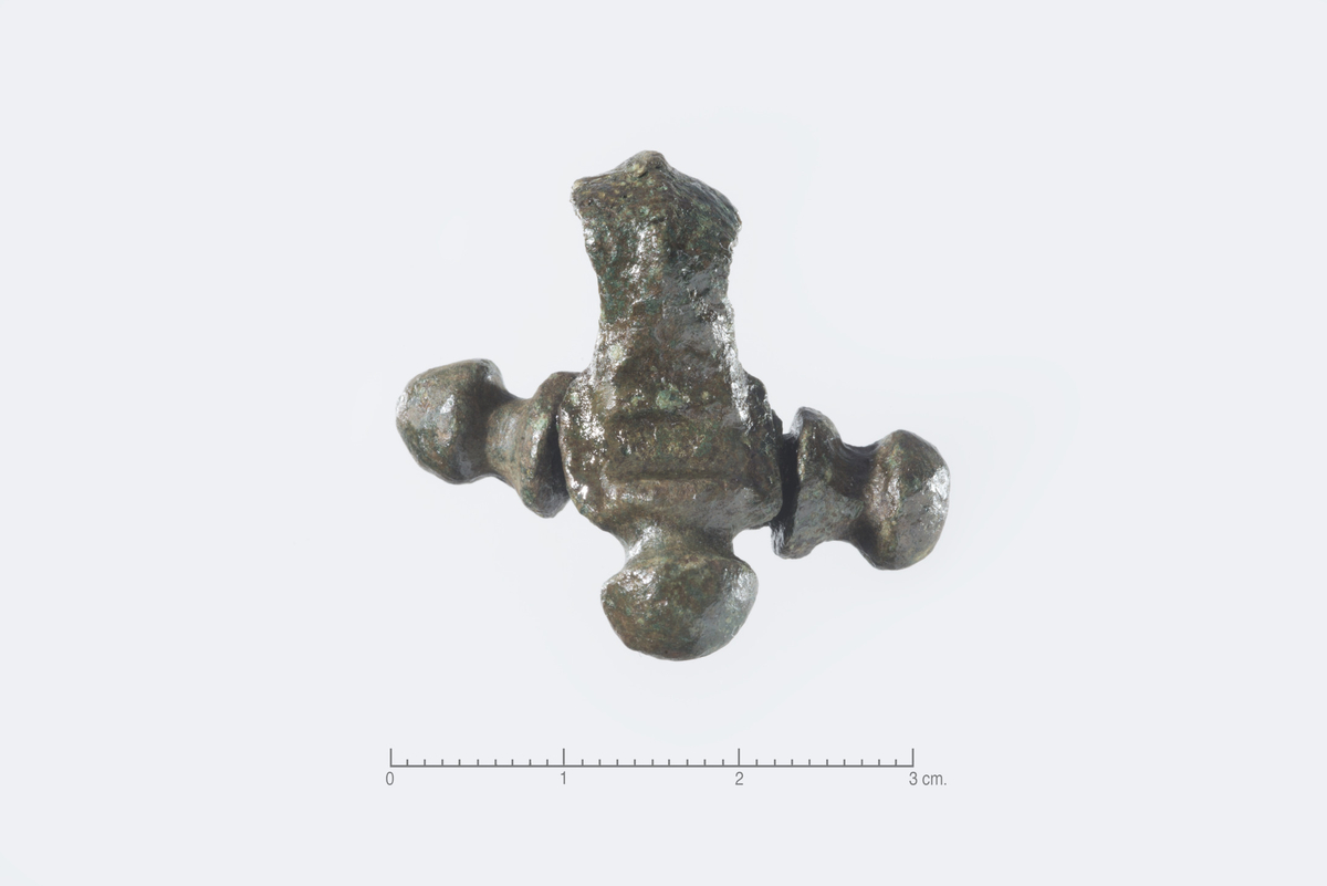Spenne, fragment av korsformet spenne av bronse, Reichsteins type Tveitane-Hunn. Spennen er lik Reichstein taf. 4 nr. 8 men platen har en rektangulær nedsenking på tvers. Nedre del av spennens fot er knekt av, en av sideknoppene er vridd, bøylen er bøyd og svakt vridd mot venstre, st.l. 29 mm.