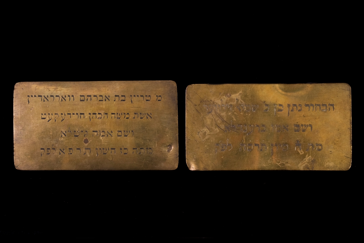 Två mässingsplattor liggandes i tygpåse med blomstermotiv. Plattorna är dekorerade med en ingraverad hebreisk text.